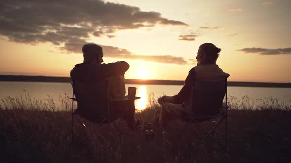 Two Men Friends Camping Near a River During Sunset Passing Food From a Parcel