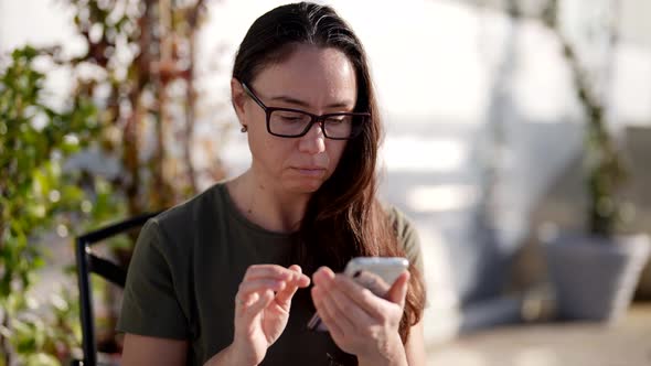 A Focused Woman with Glasses Writes a Message on Her Brand New Smartphone While Sitting at a Table