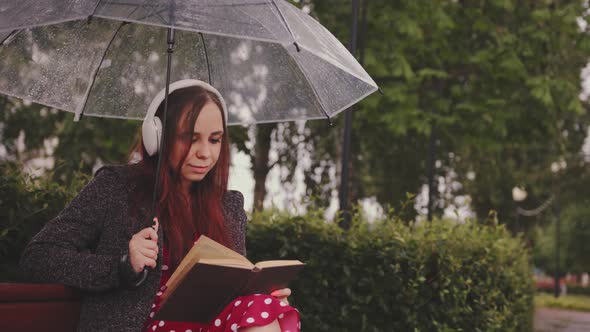 Young Beautiful Woman in Headphones with Transparent Umbrella Reading Book Sitting on Bench in City