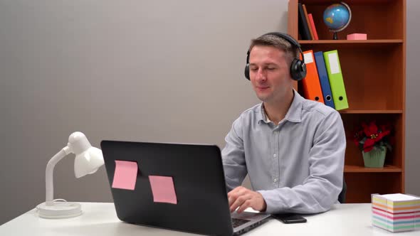 A Young Worker Communicates with a Client at an Online Conference From the Office Through Headphones