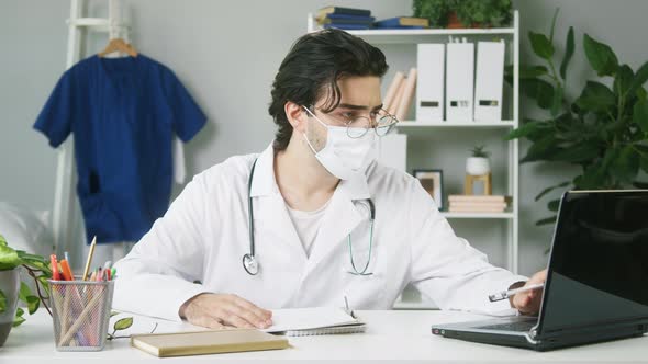 Young Doctor in Protective Mask Writing Notes Typing on Laptop Keyboard Portrait of Male Therapist