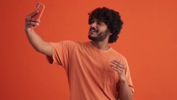 Cheerful curly-haired Indian man making selfie by phone and showing different gestures