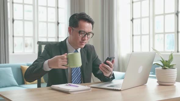 Asian Businessman Using Mobile Phone And Drinking Coffee While Using Computer Working At Home