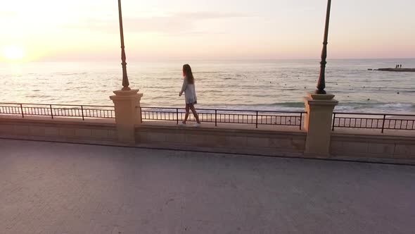 Slomo From Drone Young Woman with Long Hair in White Outfit Walking Along Parapet of Embankment