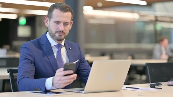 Middle Aged Businessman with Laptop Using Smartphone at Work