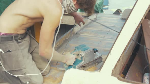 Man sands teak deck planking with belt sander on wooden boat topless in Summer sunshine.