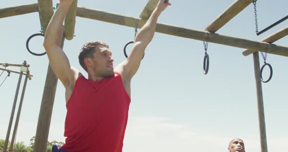 Fit caucasian man in red vest hanging from trapeze bars on obstacle course in sun