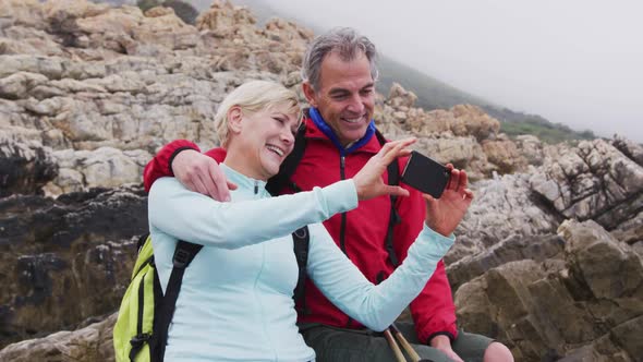 Senior hiker couple with backpacks sitting on the rocks and smiling while having a video call