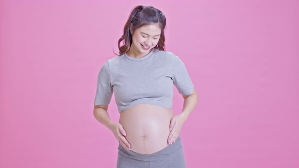 Pregnant Woman standing smile stroking big belly showing thumbs up sign isolated on pink