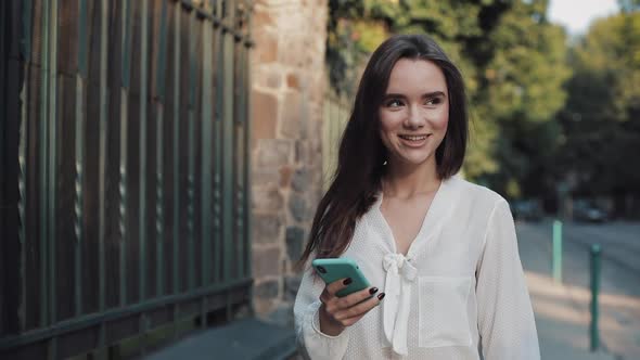 Young Attractive Smiling Brunette Girl Walking Down Cosy Street While Using Her Phone and Chatting