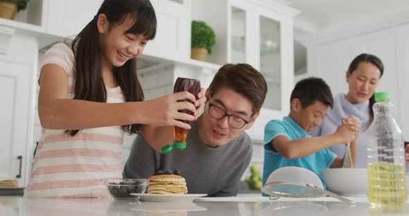 Happy asian parents in kitchen cooking with son and daughter, daughter putting syrup on pancakes