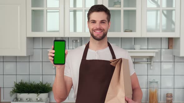 A Man in an Apron Holds a Smartphone with a Green Screen in His Hand and Smiles