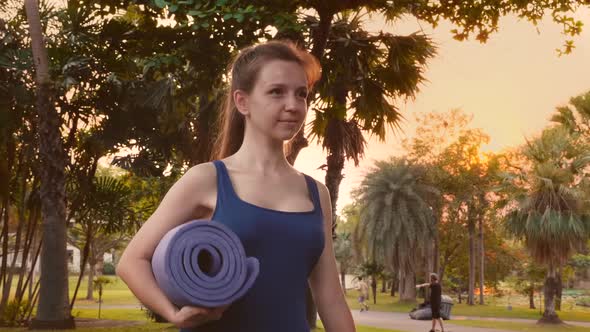 Young Fitness Woman in Sportswear Walking in City Tropical Park with Yoga Mat