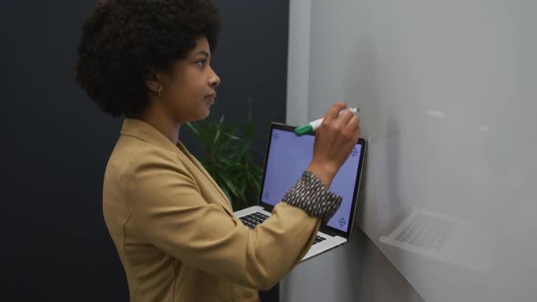 Mixed race businesswoman standing using laptop writing on a board in modern office