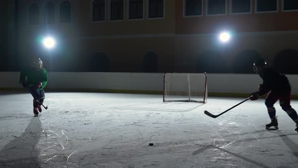 Two Men in Uniforms and Helmets with Hockey Sticks Skate on the Ice Arena Hitting Puck and Collide