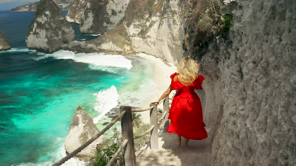 Young Woman in Red Dress Hikes Down Steep Path To Beautiful Diamond Beach in Nusa Penida Island