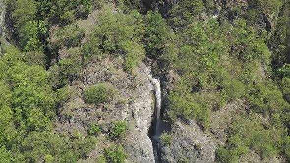 Aerial drone shot over green forest and rocky mountain cliffs with a tall waterfall in Switzerland.