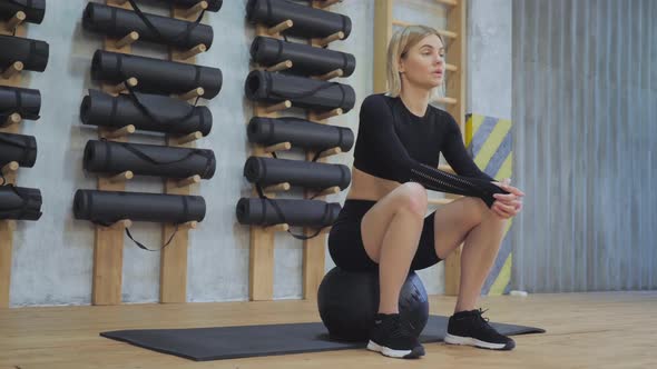 A Sporty Blonde Woman in a Black Top and Leggings Sits on the Floor on a Ball