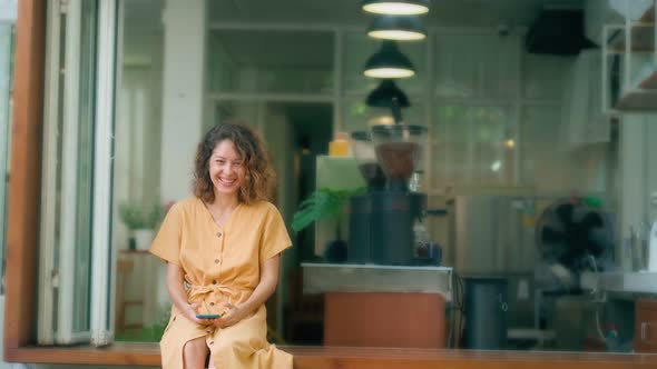 Laughing Curly Woman in Yellow Dress Using Her Phone While Sits in Coffee Shop