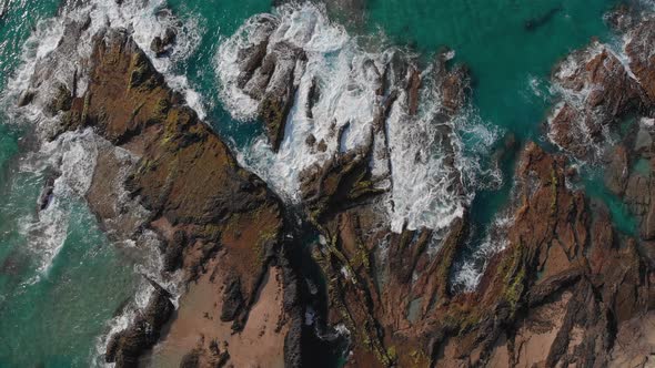 Waves hit the brown-orange rocks of the shore with their clear turquoise water turning to white foam