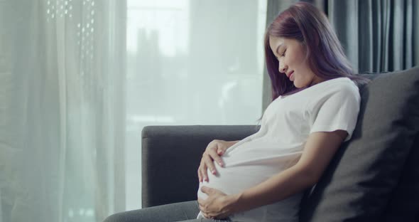 Asian pregnant woman stroking belly while sitting on a sofa in the living room at home.