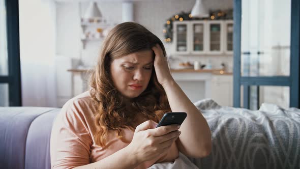 Upset Young Overweight Woman Sitting on Couch Holding Smartphone in Hands Received Message with Bad