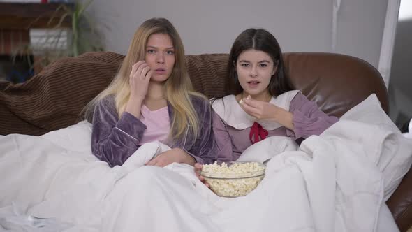 Two Absorbed Young Caucasian Women in Pajamas Eating Popcorn Looking at Camera Watching Film on TV