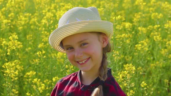 Smiling Little Girl with Hat Looking Into Camera Against Rapeseed Field