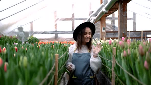 An Attractive Gardener Girl in a Hat Dances in a Good Mood in a Greenhouse Between Bright Blooming