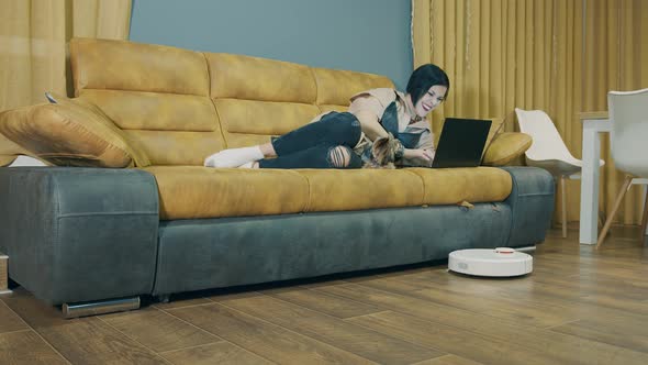 A Woman Lying on a Sofa and Working on a Laptop and a Robot Vacuum Cleaner Automatically Vacuumes