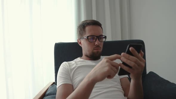 Young Man with Glasses Is Using Smartphone in Room in Daytime, Close-up View