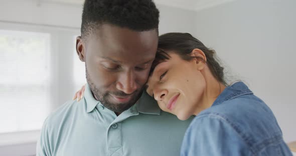 Happy diverse couple wearing jacket and shirt and embracing in living room