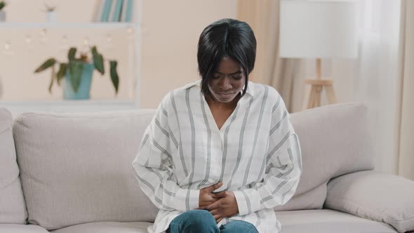 Young Upset Unhealthy Sad African American Woman Sitting in Room on Couch Holding Hands on Stomach