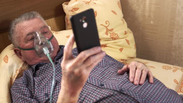 Old Sick Man is Lying on the Bed Wearing an Oxygen Mask and Watching the Mobile Phone in His Hand