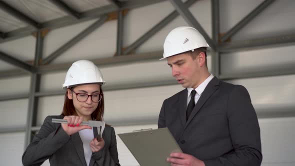 Young Man and Woman in Helmets with Documents at a Construction Site. Businessmen in Suits Measure