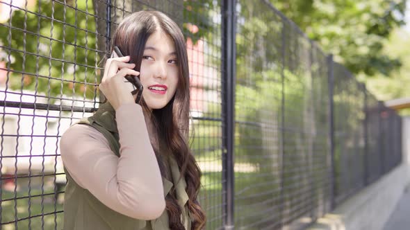 Young Asian Woman Talks Smartphone Smile As She Leans Against Tall Fence