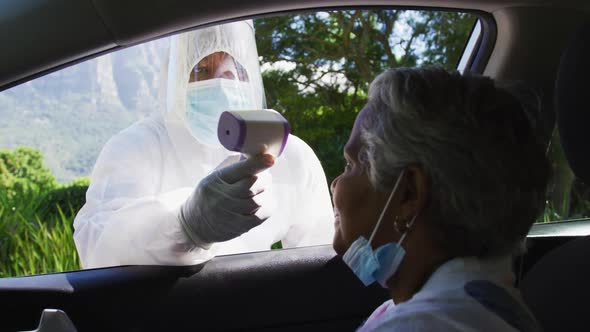 Health worker in protective clothes measuring temperature of african american senior woman in car