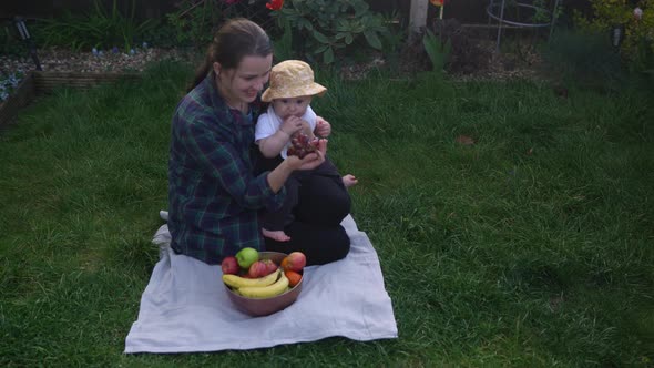 Happy Young Cheerful Mother Holding Baby Eating Fruits On Green Grass