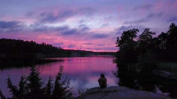 Aerial drone shot,of a man sitting at a rocky shore, of a lake, a purple sky, a colorful sunset or d