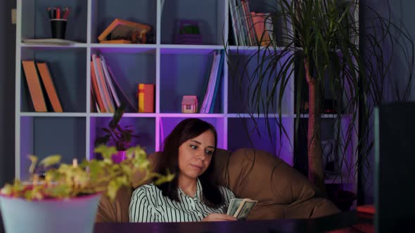 Young woman counts money, sitting in armchair before computer in office.