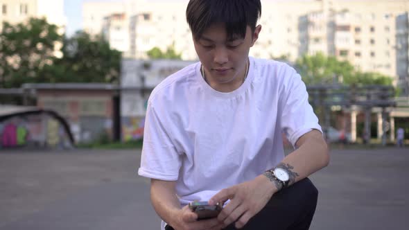 Young Asian Man Typing Message on Smartphone Outdoors