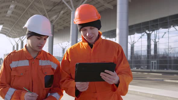 Two Construction Workers in Orange Uniform and Helmets Walking Through the Construction Field and