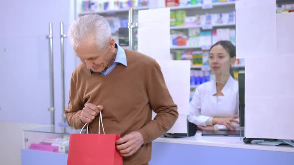 Portrait of Smiling Caucasian Greyhaired Man Posing with Shopping Bag at Cash Desk in Pharmacy