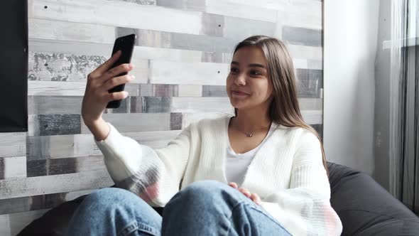 a Pretty Young Girl in a Light Jacket is Sitting on a Chair at Home and Talking Via Video Link Via a