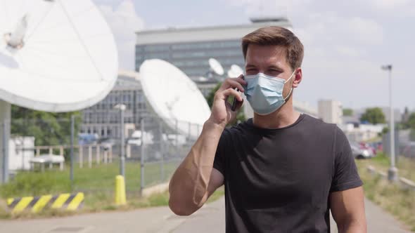 A Young Handsome Man in a Face Mask Talks on a Smartphone in an Urban Area - Satellite Dishes