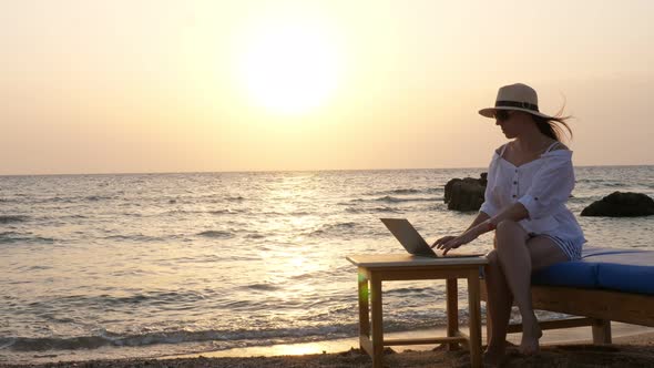 Young Woman in Sunglasses and Sun Hat, Uses Laptop, Sitting on the Beach By the Sea, at Sunset or