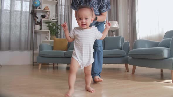 Asian Father Encouraging Smiling Baby Son To Take First Steps And Walk At Home