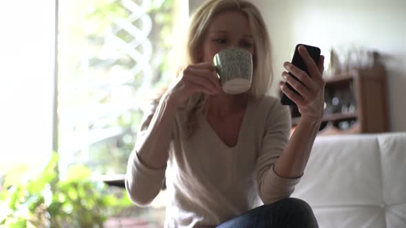 Mature woman drinking coffee and using smart phone in living room