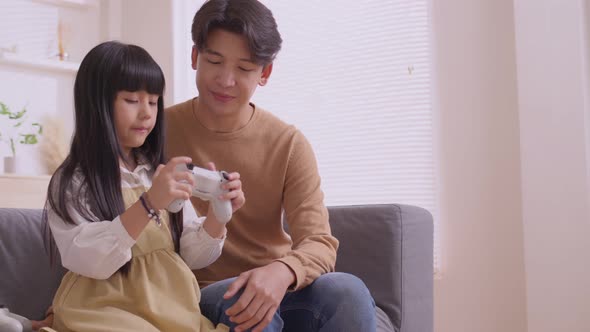 Happy Asian father teaching daughter to play games with joy stick on sofa in relax day.