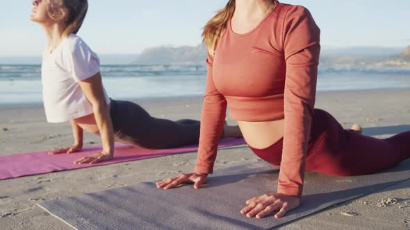 Two caucasian female friends practicing yoga, stretching at the beach